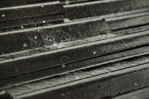 Raindrops splash on a dark metal roof, creating small ripples and droplets. The surface is textured, with water glistening in a dimly lit environment.