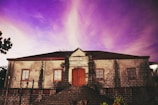 A historic stone building with a central wooden door and two windows on each side. The structure has a reddish-brown roof and is set against a vibrant, purple-hued sky. A sign above the door reads 'Taal Cultural Center'. Stairs lead up to the entrance, and some vegetation is visible at the foreground.