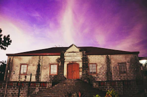 A historic stone building with a central wooden door and two windows on each side. The structure has a reddish-brown roof and is set against a vibrant, purple-hued sky. A sign above the door reads 'Taal Cultural Center'. Stairs lead up to the entrance, and some vegetation is visible at the foreground.