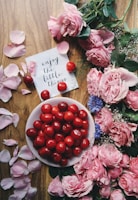A wooden surface decorated with pink flowers and petals on the right side and around the edges. In the center, there's a white plate filled with bright red cherries. To the left of the plate, there is a handwritten note saying 'enjoy the little things' with a few cherries scattered around.