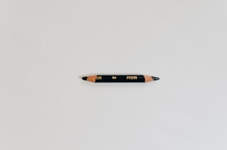A black colored pencil with sharpened ends, placed horizontally on a light gray background. The pencil has golden inscribed text and wooden tips that show the pencil lead.
