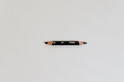 A black colored pencil with sharpened ends, placed horizontally on a light gray background. The pencil has golden inscribed text and wooden tips that show the pencil lead.