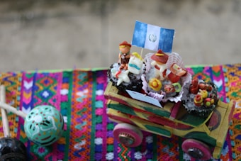 A wooden toy truck is decorated with various small, colorful figures made of candy or dough, dressed in traditional attire and positioned on top. A small flag is also present among the figures. The truck is resting on a vibrantly colored fabric featuring intricate designs.