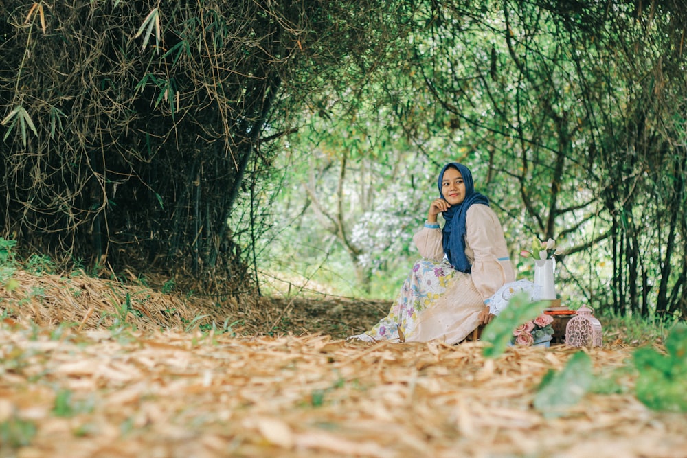 A woman in a blue headscarf and a floral dress smiles while sitting under a green arch