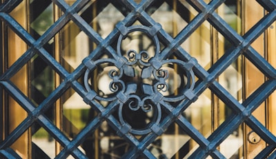 An intricate wrought iron grille with a decorative floral pattern is seen in a close-up view. The grille features diagonal lines forming diamond shapes, and a central ornate design with scrollwork. Behind the grille, there is a glimpse of a reflective golden surface, possibly a door or window.