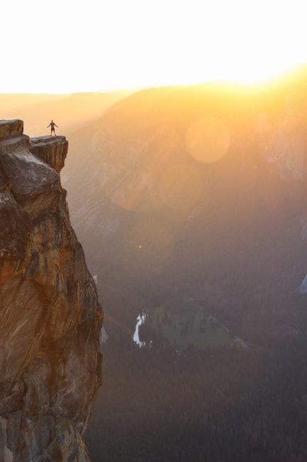A lone figure stands triumphantly on the edge of a high cliff, overlooking a vast valley bathed in warm sunlight. The sun is setting or rising, casting a golden hue over the scene and creating a dramatic lens flare effect. The cliff's rugged texture and the expansive view of the valley contribute to a sense of adventure and solitude.