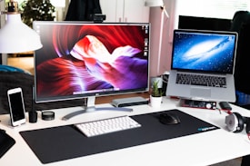 A modern desk setup featuring a large curved monitor displaying vibrant abstract art. Next to it is an open laptop with a galaxy wallpaper. The desk holds a smartphone, a mouse, a keyboard, a small potted plant, and headphones. A desk lamp provides additional lighting, while various small gadgets and a black desk mat complete the arrangement.