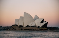 A famous opera house with a series of white sail-like shells on its roof, situated against a backdrop of an evening sky transitioning from pink to blue. The structure is bordered by water, and the distant skyline and waterfront create a serene urban scene.
