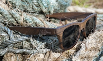 A pair of wooden sunglasses is resting on a pile of thick, fibrous ropes with a mix of green and beige tones. The sunglasses have dark lenses and the wood grain is clearly visible on the frames.