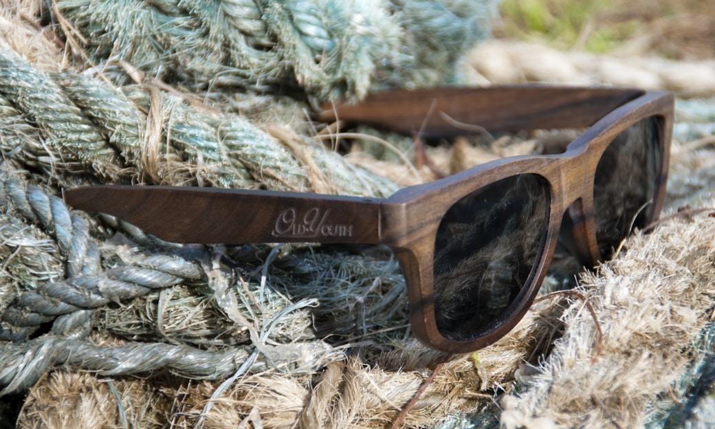 A pair of wooden sunglasses is resting on a pile of thick, fibrous ropes with a mix of green and beige tones. The sunglasses have dark lenses and the wood grain is clearly visible on the frames.