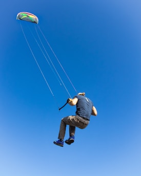 A person is engaged in kitesurfing against a clear blue sky. The kite is multi-colored and is connected to the person with several lines. The individual is airborne, wearing casual outdoor clothing, and appears to be enjoying the activity.
