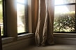 Curtains are gently draped at the corner of a living room window overlooking some greenery and a parked car outside. The sunlight streams in through the clear glass, casting a warm glow on the neutral-colored fabric.