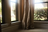 Curtains are gently draped at the corner of a living room window overlooking some greenery and a parked car outside. The sunlight streams in through the clear glass, casting a warm glow on the neutral-colored fabric.