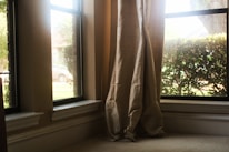 Curtains are gently draped at the corner of a living room window overlooking some greenery and a parked car outside. The sunlight streams in through the clear glass, casting a warm glow on the neutral-colored fabric.