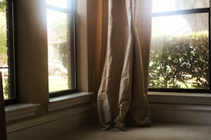 Curtains are gently draped at the corner of a living room window overlooking some greenery and a parked car outside. The sunlight streams in through the clear glass, casting a warm glow on the neutral-colored fabric.