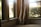 Curtains are gently draped at the corner of a living room window overlooking some greenery and a parked car outside. The sunlight streams in through the clear glass, casting a warm glow on the neutral-colored fabric.