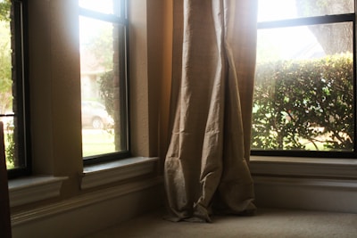 Curtains are gently draped at the corner of a living room window overlooking some greenery and a parked car outside. The sunlight streams in through the clear glass, casting a warm glow on the neutral-colored fabric.