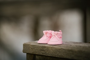 A pair of tiny pink baby shoes with bows and a soft fabric design resting on a weathered wooden surface. The background is softly blurred, giving a shallow depth of field effect.