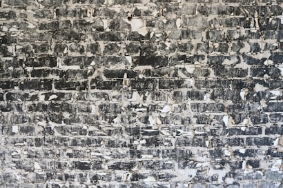 A close-up of a weathered brick wall with peeling paint and a rough texture. The bricks are unevenly covered with black and white paint patches, giving a distressed appearance.