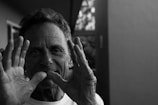 An older person smiles while holding up their hands near their face, with the image captured in black and white. The background appears to show the interior of a home and an open window.