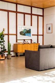 A modern living room with wooden paneling and a large framed artwork hanging on the wall above a mid-century style wooden credenza. The room features a gray sofa in the foreground, minimal decor with two green potted plants and a brass floor lamp to the left. The floor is polished concrete, and the space feels airy and light-filled with a clean, minimalist aesthetic.