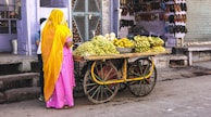 A woman dressed in a pink sari with a yellow head covering stands beside a wooden cart filled with fresh fruits such as grapes, bananas, and oranges. The cart has large wooden wheels and is situated on a street in front of a shop displaying an array of footwear. The background has purple-painted walls and a closed metal shutter on one side.