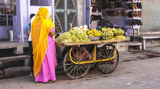 A woman dressed in a pink sari with a yellow head covering stands beside a wooden cart filled with fresh fruits such as grapes, bananas, and oranges. The cart has large wooden wheels and is situated on a street in front of a shop displaying an array of footwear. The background has purple-painted walls and a closed metal shutter on one side.