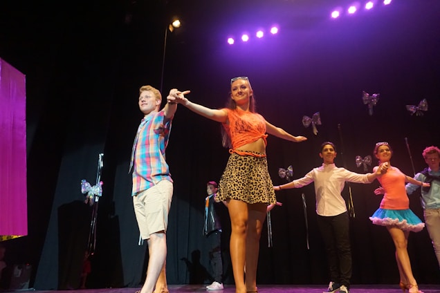 A group of performers on stage under bright stage lights. The central figures include a man in a plaid shirt and khaki shorts and a woman in a pinkish-orange top and leopard print skirt. Additional performers in various colorful outfits can be seen in the background, with large decorative bows as part of the stage design.