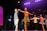A group of performers on stage under bright stage lights. The central figures include a man in a plaid shirt and khaki shorts and a woman in a pinkish-orange top and leopard print skirt. Additional performers in various colorful outfits can be seen in the background, with large decorative bows as part of the stage design.