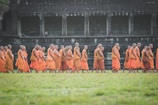 A group of Buddhist monks dressed in vibrant orange robes walk in a procession across a lush green field. The backdrop features an ancient stone structure with arches, showing signs of historical architecture and wear.