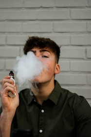A person wearing a dark green button-up shirt exhales vapor while holding a vaping device. The background consists of a light gray brick wall.