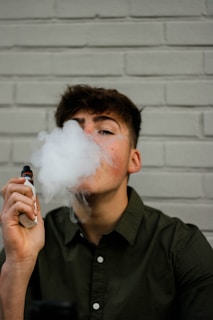 A person wearing a dark green button-up shirt exhales vapor while holding a vaping device. The background consists of a light gray brick wall.
