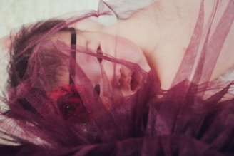 A sleeping baby partially covered with a soft, sheer burgundy fabric. The fabric gently drapes across the baby’s face and forehead, creating a serene and peaceful atmosphere.