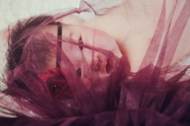 A sleeping baby partially covered with a soft, sheer burgundy fabric. The fabric gently drapes across the baby’s face and forehead, creating a serene and peaceful atmosphere.