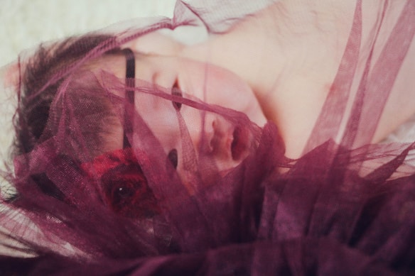 A sleeping baby partially covered with a soft, sheer burgundy fabric. The fabric gently drapes across the baby&rsquo;s face and forehead, creating a serene and peaceful atmosphere.