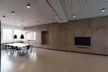 A contemporary interior space featuring a minimalist design with wooden wall panels, a long table surrounded by black chairs, and pendant lights hanging from the ceiling. The room is lit with natural light from large windows, and bench seating is integrated into the walls. A large flat-screen TV is mounted on the wall, contributing to the modern aesthetic.