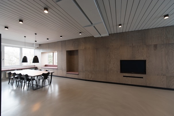 A contemporary interior space featuring a minimalist design with wooden wall panels, a long table surrounded by black chairs, and pendant lights hanging from the ceiling. The room is lit with natural light from large windows, and bench seating is integrated into the walls. A large flat-screen TV is mounted on the wall, contributing to the modern aesthetic.