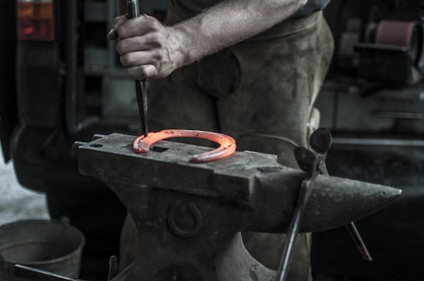A blacksmith is in the process of forging a horseshoe, which is glowing orange from the heat. The worker's hands are holding a tool and hammering on an anvil, with other blacksmithing tools visible in the background.