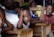 A classroom setting with several children seated at wooden desks. One child in the foreground rests their head on their hand, gazing directly ahead, while others around them are smiling and engaged.