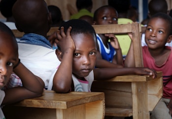 A classroom setting with several children seated at wooden desks. One child in the foreground rests their head on their hand, gazing directly ahead, while others around them are smiling and engaged.