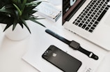 A neatly arranged workspace featuring a laptop with a partial view of its keyboard and screen displaying time. Nearby, a black smartwatch and a black smartphone rest on a white surface, accompanied by a small stack of magazines. A green plant in a white pot adds a touch of nature.
