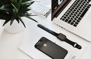 A neatly arranged workspace featuring a laptop with a partial view of its keyboard and screen displaying time. Nearby, a black smartwatch and a black smartphone rest on a white surface, accompanied by a small stack of magazines. A green plant in a white pot adds a touch of nature.