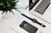 A neatly arranged workspace featuring a laptop with a partial view of its keyboard and screen displaying time. Nearby, a black smartwatch and a black smartphone rest on a white surface, accompanied by a small stack of magazines. A green plant in a white pot adds a touch of nature.