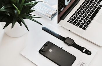 A neatly arranged workspace featuring a laptop with a partial view of its keyboard and screen displaying time. Nearby, a black smartwatch and a black smartphone rest on a white surface, accompanied by a small stack of magazines. A green plant in a white pot adds a touch of nature.