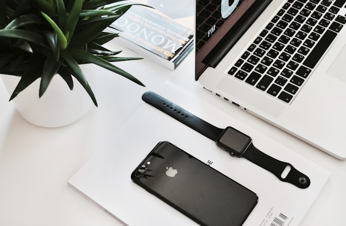 A neatly arranged workspace featuring a laptop with a partial view of its keyboard and screen displaying time. Nearby, a black smartwatch and a black smartphone rest on a white surface, accompanied by a small stack of magazines. A green plant in a white pot adds a touch of nature.
