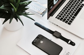 A neatly arranged workspace featuring a laptop with a partial view of its keyboard and screen displaying time. Nearby, a black smartwatch and a black smartphone rest on a white surface, accompanied by a small stack of magazines. A green plant in a white pot adds a touch of nature.
