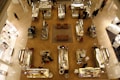 An overhead view of a high-end department store showcasing several cosmetic and jewelry counters arranged in an organized grid pattern on a polished wooden floor. Each counter is attended by staff assisting customers, with displays of various luxury goods. There are brown leather chairs placed in the central area, providing a place for rest or consultation.
