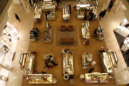 An overhead view of a high-end department store showcasing several cosmetic and jewelry counters arranged in an organized grid pattern on a polished wooden floor. Each counter is attended by staff assisting customers, with displays of various luxury goods. There are brown leather chairs placed in the central area, providing a place for rest or consultation.