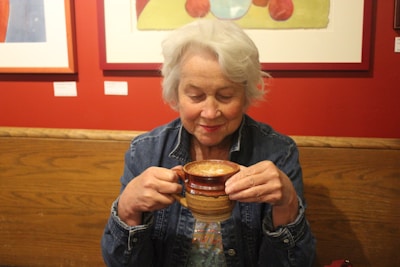 An older woman with white hair enjoys a hot beverage in a ceramic mug. She is wearing a denim jacket, and there are colorful paintings on a red wall in the background. She appears content and relaxed, sitting in a cozy café environment.