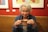An older woman with white hair enjoys a hot beverage in a ceramic mug. She is wearing a denim jacket, and there are colorful paintings on a red wall in the background. She appears content and relaxed, sitting in a cozy café environment.